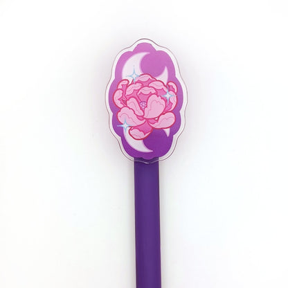 A gel pen with an acrylic charm featuring a peony design. The flower is pink on a purple backdrop, with two moons behind it