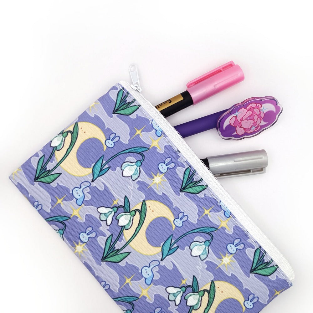 A pencil case with three pens sticking out. It is purple with a smoky background. It features stars, the moon, snowdrops, and cute bunny heads floating around.