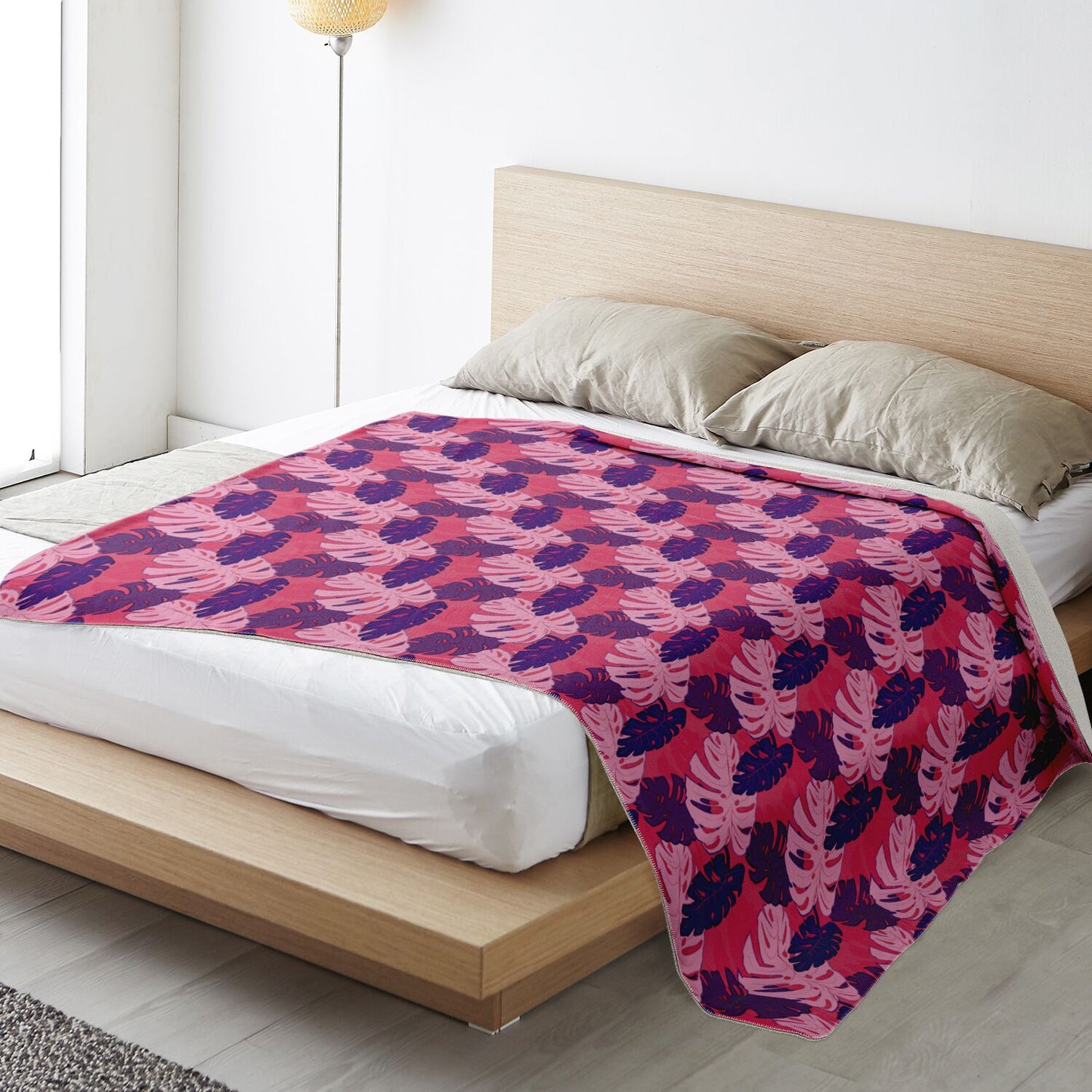 A blanket draped over a bed. The blanket has a monstera pattern with purple and light pink leaves on a pink background.