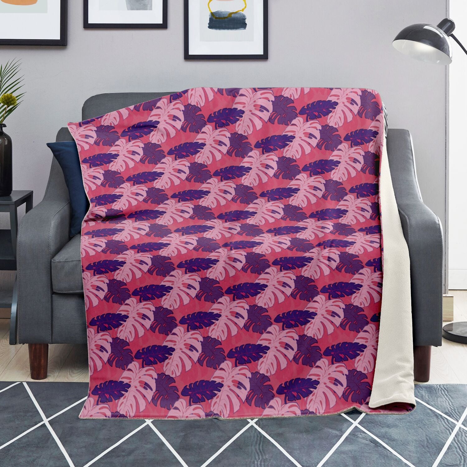 A blanket draped over a couch. The blanket has a monstera pattern with purple and light pink leaves on a pink background.