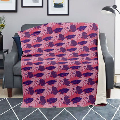 A blanket draped over a couch. The blanket has a monstera pattern with purple and light pink leaves on a pink background.