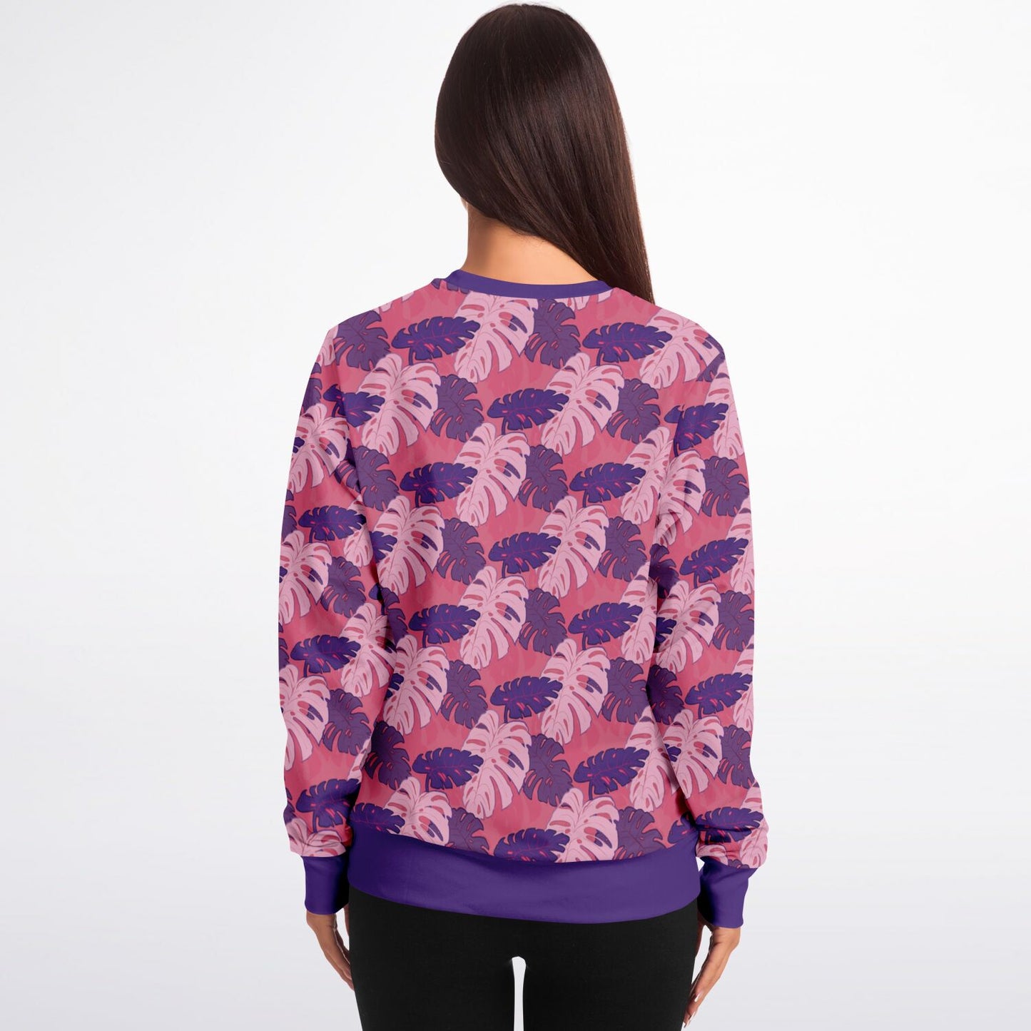 A model wearing a sweatshirt. The sweatshirt's pattern features monstera leaves with purple and light pink leaves on a pink background.