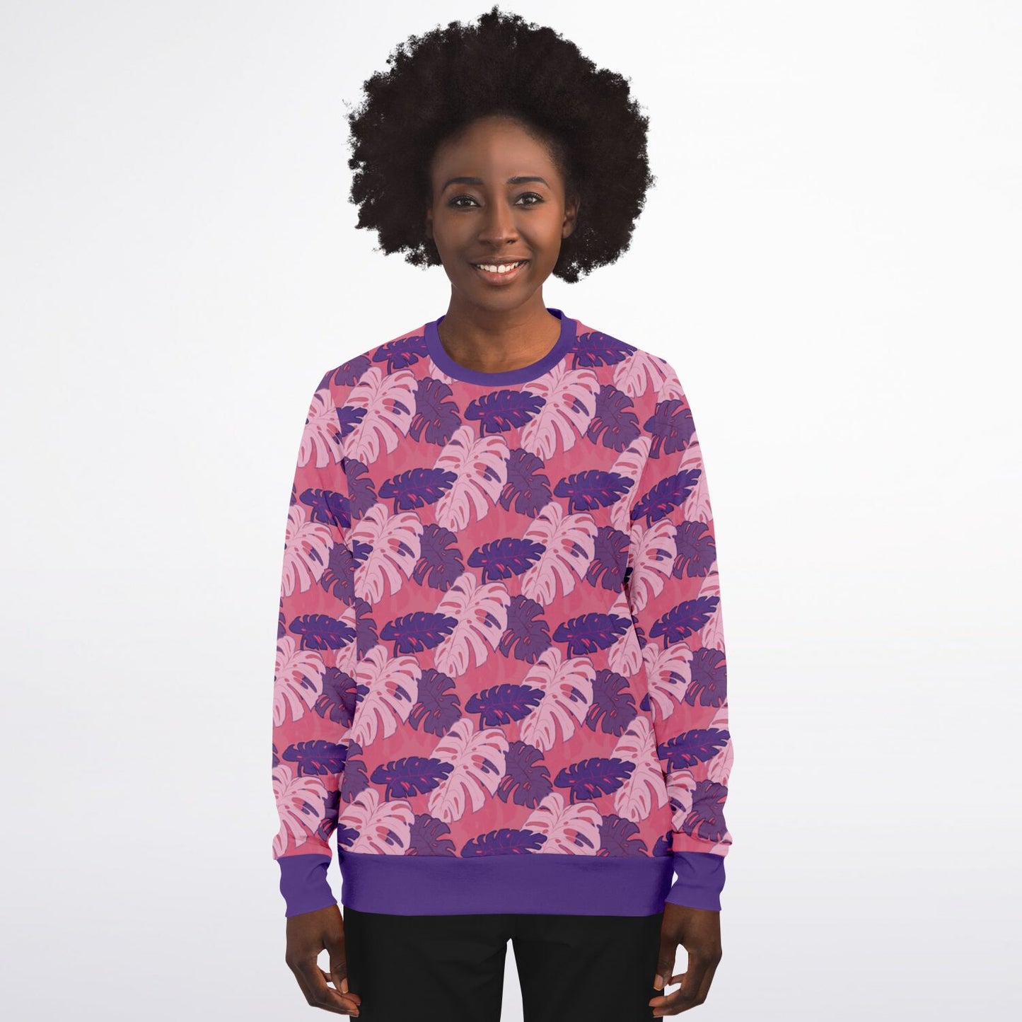 A model wearing a sweatshirt. The sweatshirt's pattern features monstera leaves with purple and light pink leaves on a pink background.