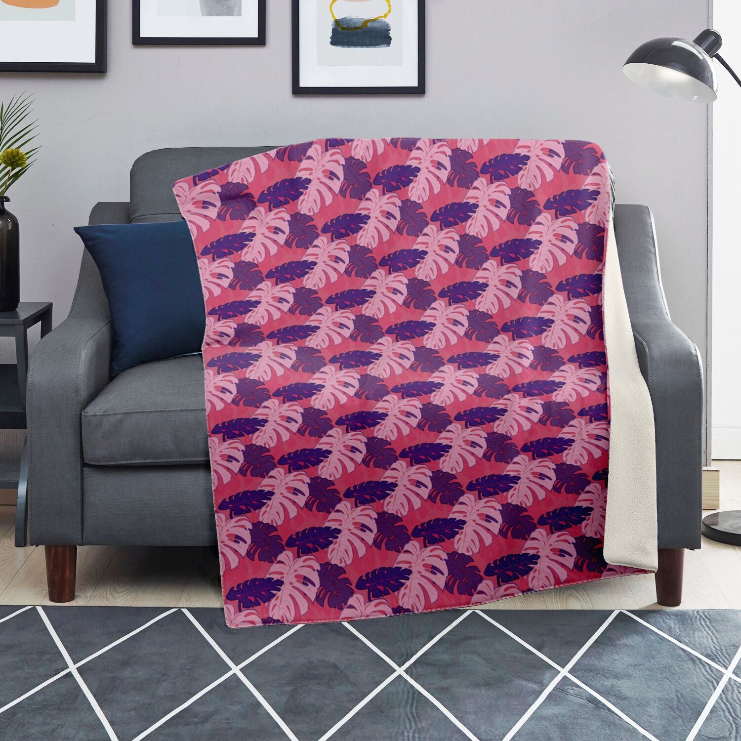 A blanket draped over a couch. The blanket has a monstera pattern with purple and light pink leaves on a pink background.