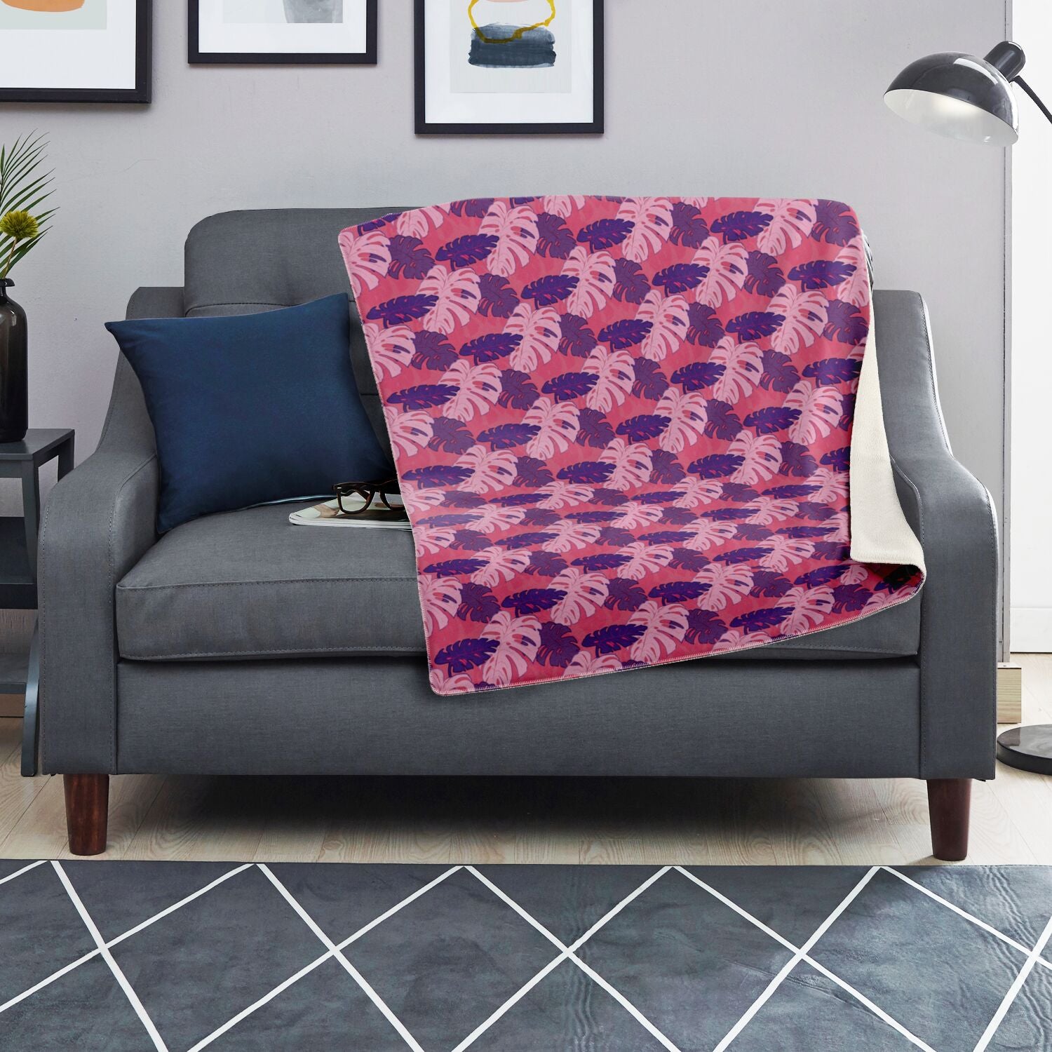 A blanket draped over a couch. The blanket has a monstera pattern with purple and light pink leaves on a pink background.